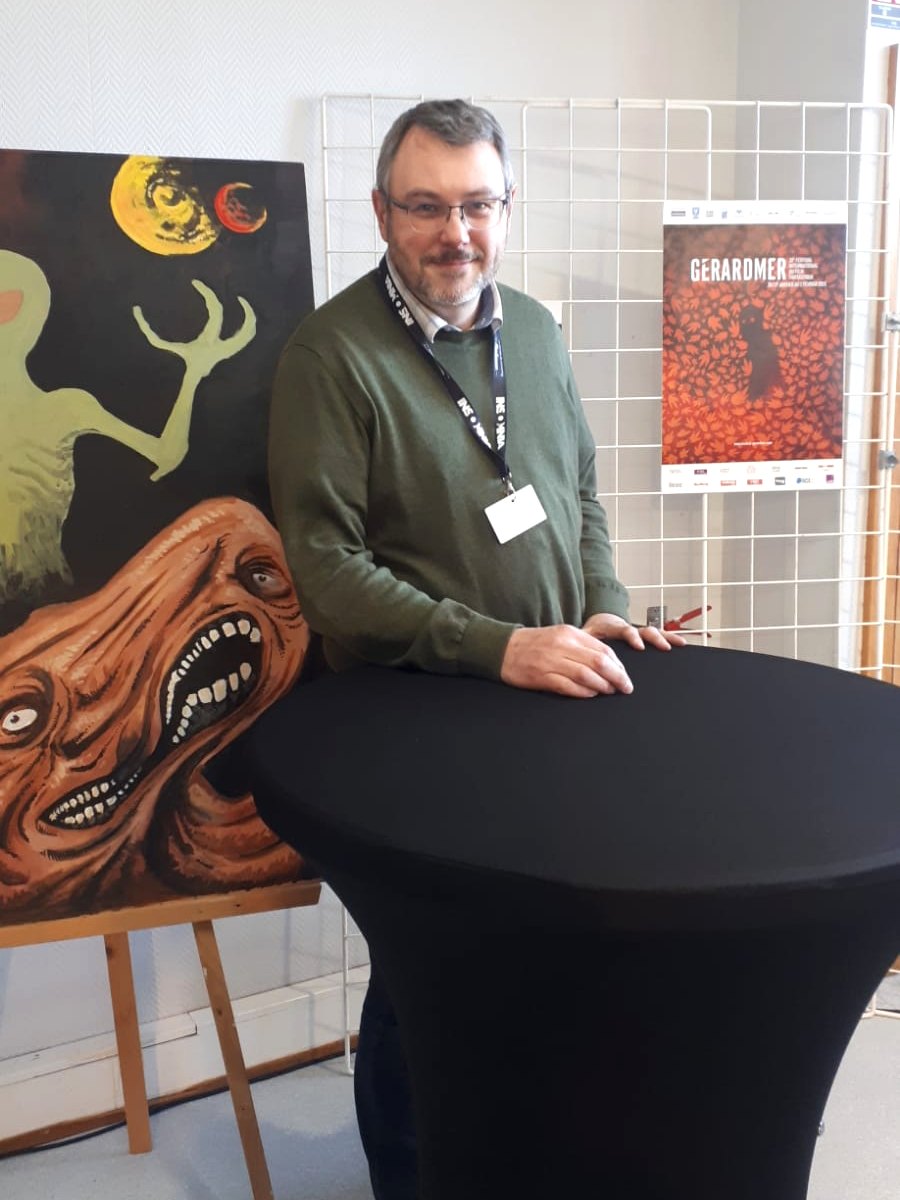 Jean K. Saintfort devant un tableau de monstre et l’affiche du 32e Festival de Gérardmer